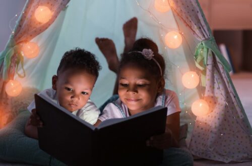 a little boy and little girl reading a children's book in a tent at night surrounded by white fairy lights